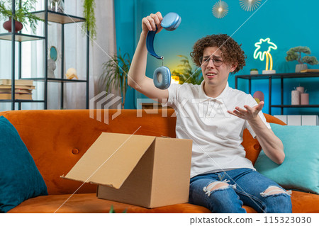 Unhappy man unpacking carton box feeling dissatisfied with getting damaged wireless headphones home Unhappy man unpacking carton box feeling dissatisfied with getting damaged wireless headphones home 115323030