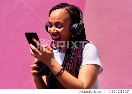 Woman, headphones and happy music for podcast in the city, pink wall background and call on smartphone. Streaming, funny and female with coffee listening to track, singing on cellphone and excited 115323507