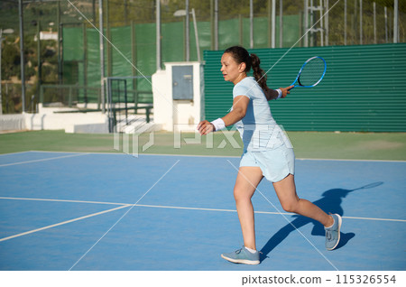 Middle-aged female tennis player hitting a backhand shot on outdoor court 115326554