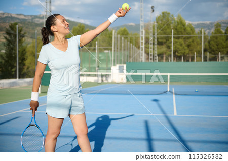 Middle-aged woman playing tennis on an outdoor court, preparing to serve 115326582