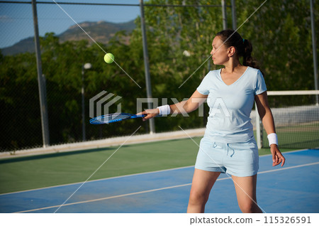 Middle-aged woman playing tennis on outdoor court under bright sunlight Middle-aged woman playing tennis on outdoor court under bright sunlight 115326591