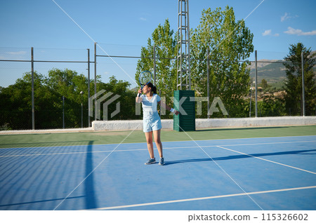 Middle-aged woman playing tennis on outdoor court under bright sunny sky 115326602