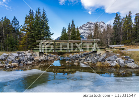 Wooden footbridge in the forest across frozen icy water surface in winter. Snow-covered mountain in background. Johnson lake, Banff National Park, Canadian Rockies, Alberta, Canada. Wooden footbridge in the forest across frozen icy water surface in winter. Snow-covered mountain in background. Johnson lake, Banff National Park, Canadian Rockies, Alberta, Canada. 115331547