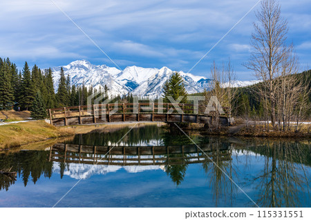 Wooden arch footbridge in Cascade Ponds park in autumn sunny day, snow-covered Mount Astley reflection on the water surface. Banff National Park, Canadian Rockies, Alberta, Canada. 115331551