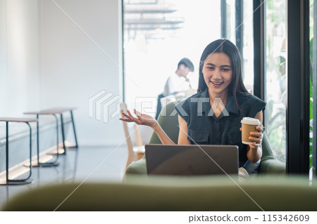 A woman is sitting in a chair with a laptop in front of her. She is holding a cup of coffee and she is talking on the phone. The scene suggests a casual and relaxed atmosphere A woman is sitting in a chair with a laptop in front of her. She is holding a cup of coffee and she is talking on the phone. The scene suggests a casual and relaxed atmosphere 115342609