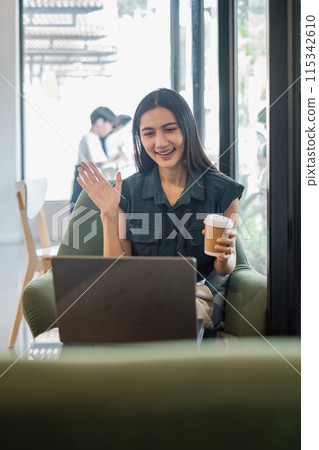 A woman is sitting in a chair with a laptop in front of her. She is smiling and waving at the camera. She is holding a cup of coffee A woman is sitting in a chair with a laptop in front of her. She is smiling and waving at the camera. She is holding a cup of coffee 115342610