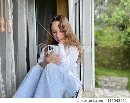 a girl in a white shirt a teenager sits on a white windowsill the view from the street she can be seen in the window in her hands she holds a white iPhone 13 pro max she looks at the screen calmly a girl in a white shirt a teenager sits on a white windowsill the view from the street she can be seen in the window in her hands she holds a white iPhone 13 pro max she looks at the screen calmly 115342831