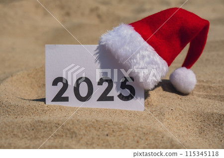 2025 text on paper greeting card on background sandy beach sun coast. Christmas Santa hat New Year New Me decoration. Summer vacation decor. Holiday concept calendar date postcard. Getting away 115345118