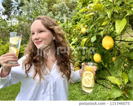 it's just a lemon paradise, a teenage girl stands against the backdrop of a lemon tree with lemons and drinks lemonade from a glass glass in her other hand, she holds a jug of drink 115346075
