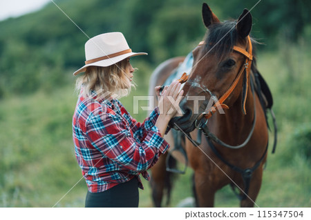 Happy blonde with horse in forest. Woman and a horse walking through the field during the day. Dressed in a plaid shirt and black leggings. Happy blonde with horse in forest. Woman and a horse walking through the field during the day. Dressed in a plaid shirt and black leggings. 115347504