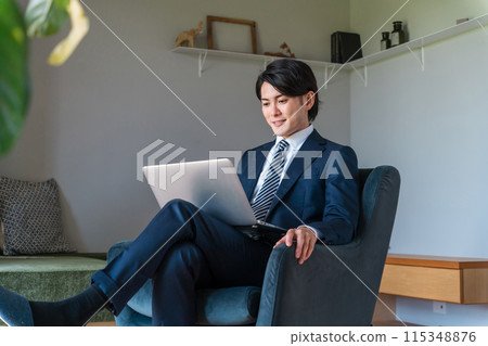 A young Asian businessman in a suit relaxing on a sofa looking at a laptop A young Asian businessman in a suit relaxing on a sofa looking at a laptop 115348876