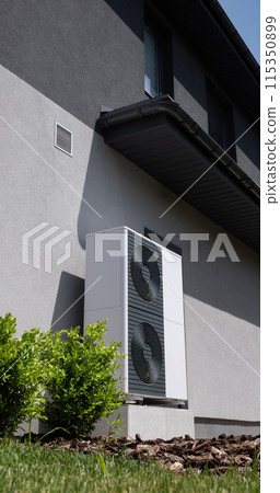 The outdoor unit of a heat pump is mounted against the wall of a modern cottage. The outdoor unit of a heat pump is mounted against the wall of a modern cottage. 115350899