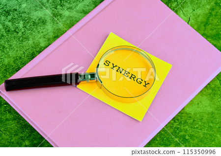 Business concept Synergy word through a magnifying glass on a yellow sticker lying on a business notebook on a green background 115350996