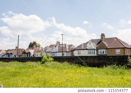A row of houses with triangular roofs and a field of yellow flowers blooming under bright sunlight with white clouds floating in the sky, in the suburbs of London A row of houses with triangular roofs and a field of yellow flowers blooming under bright sunlight with white clouds floating in the sky, in the suburbs of London 115354377