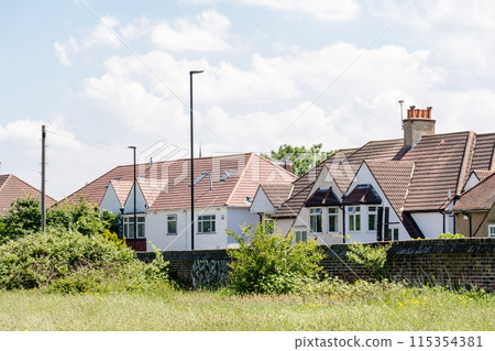 A row of houses with triangular roofs and a field of yellow flowers blooming under bright sunlight with white clouds floating in the sky, in the suburbs of London 115354381