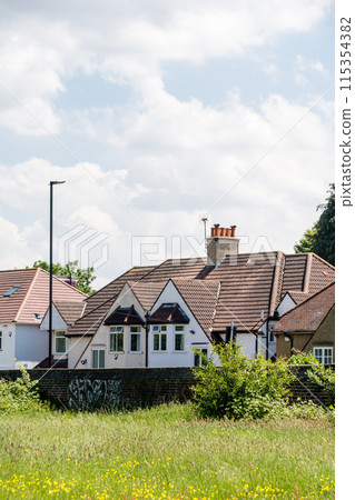 A row of houses with triangular roofs and a field of yellow flowers blooming under bright sunlight with white clouds floating in the sky, in the suburbs of London A row of houses with triangular roofs and a field of yellow flowers blooming under bright sunlight with white clouds floating in the sky, in the suburbs of London 115354382