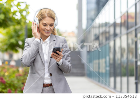 Smiling businesswoman in gray suit using a smartphone and listening to music with headphones. Modern urban background. Smiling businesswoman in gray suit using a smartphone and listening to music with headphones. Modern urban background. 115354518