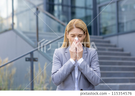 Businesswoman sneezing outside office building due to seasonal allergies. Professional woman suffering from cold or flu symptoms 115354576