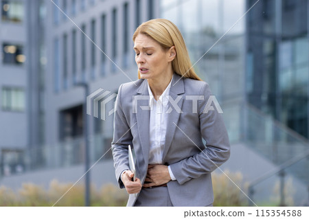 Blonde businesswoman in gray suit holding her stomach, showing discomfort and pain, standing outside modern office building in city. Blonde businesswoman in gray suit holding her stomach, showing discomfort and pain, standing outside modern office building in city. 115354588