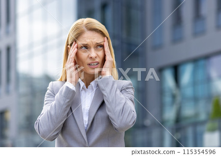 Businesswoman experiencing stress and holding her head in frustration outside a modern office building. Concept of workplace anxiety, pressure, and professional stress. 115354596