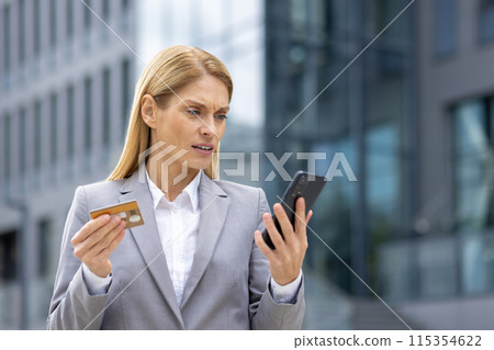 Businesswoman with a worried expression holding a credit card and smartphone. She is standing outside an office building, suspecting possible fraud or financial issue. Businesswoman with a worried expression holding a credit card and smartphone. She is standing outside an office building, suspecting possible fraud or financial issue. 115354622