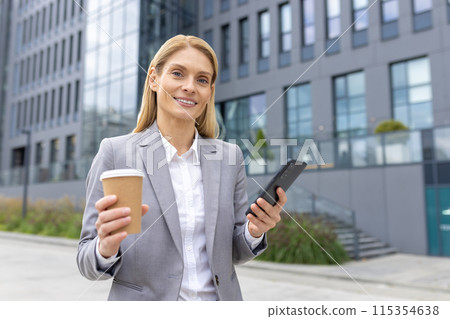 Confident businesswoman in a gray suit holding coffee and smartphone outside a modern office building. Confident businesswoman in a gray suit holding coffee and smartphone outside a modern office building. 115354638