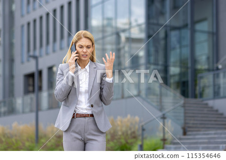 Businesswoman in a suit expressing frustration while talking on the phone outside an office building. Professional communication challenge concept. Businesswoman in a suit expressing frustration while talking on the phone outside an office building. Professional communication challenge concept. 115354646