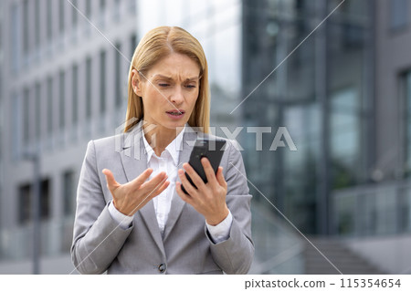 A professional woman looks upset while reading bad news on her phone. She stands outside a modern commercial building, wearing a grey suit. 115354654