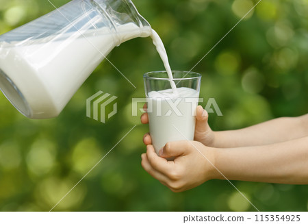 child hands holding glass and milk pouring from jug with green garden on the background 115354925
