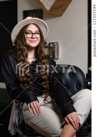 Portrait of a young hairdresser girl in a hat. A female barber is sitting in a chair and waiting for a client. 115355594
