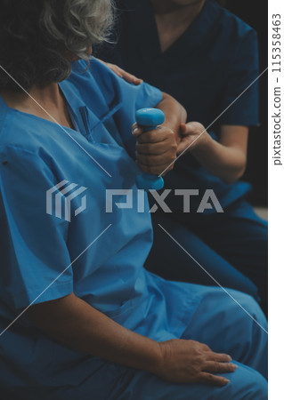 Old woman training with physiotherapist using dumbbells at home. Therapist assisting senior woman with exercises in nursing home. Elderly patient using dumbbells with outstretched arms. 115358463