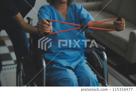Personal trainer assisting senior woman with resistance band. Rehabilitation physiotherapy worker helping old patient at nursing home. Old woman with stretch band being coached by physiotherapist. Personal trainer assisting senior woman with resistance band. Rehabilitation physiotherapy worker helping old patient at nursing home. Old woman with stretch band being coached by physiotherapist. 115358477