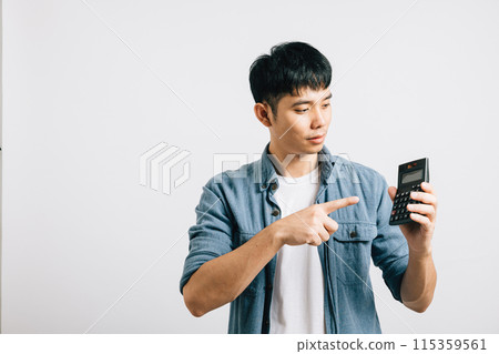 A young man, isolated on grey, points at a calculator number, conveying the success of tax calculation. His smile reflects financial achievement and career progress. A young man, isolated on grey, points at a calculator number, conveying the success of tax calculation. His smile reflects financial achievement and career progress. 115359561
