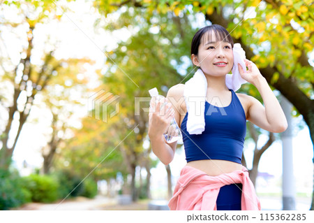 A young woman jogging along a road lined with autumn trees; dieting, body shaping; wiping sweat with a towel A young woman jogging along a road lined with autumn trees; dieting, body shaping; wiping sweat with a towel 115362285