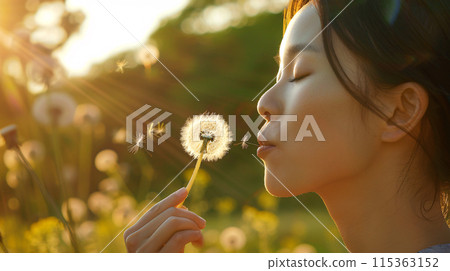 woman with a dreamy expression, holding a single dandelion seed and blowing it gently into the air 115363152