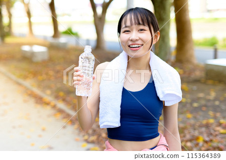 Young woman walking along a road lined with autumn trees, diet, body makeover, hydration with a plastic bottle, trimming 115364389
