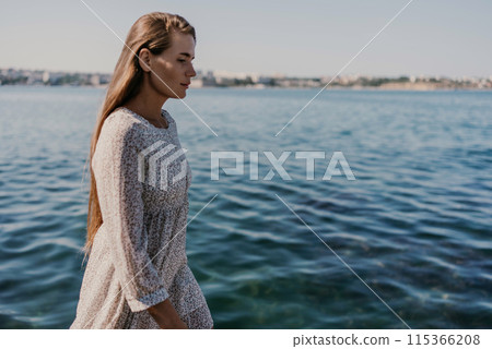 A woman is walking on the beach wearing a dress. The water is calm and the sky is clear. 115366208