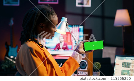 African american girl holds phone with isolated greenscreen at home, studying economics and finances at university. Young woman uses blank mockup template while she works on spreadsheets. Camera A. African american girl holds phone with isolated greenscreen at home, studying economics and finances at university. Young woman uses blank mockup template while she works on spreadsheets. Camera A. 115366290
