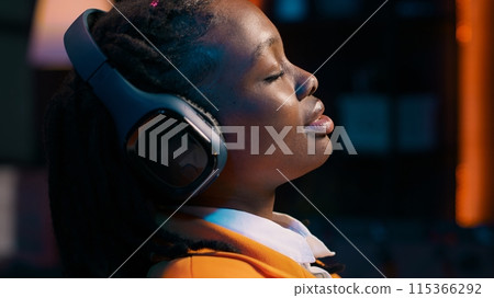 College student sitting laid back after working on class project, relaxing with ambience music on headphones at home desk. African american girl chilling with modern tunes on headset. Camera A. 115366292
