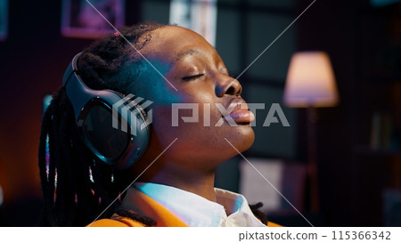 Carefree student finishes creating a presentation and relaxes with music on audio headset, resting after long researching session. African american girl chilling with ambience tunes. Camera B. Carefree student finishes creating a presentation and relaxes with music on audio headset, resting after long researching session. African american girl chilling with ambience tunes. Camera B. 115366342