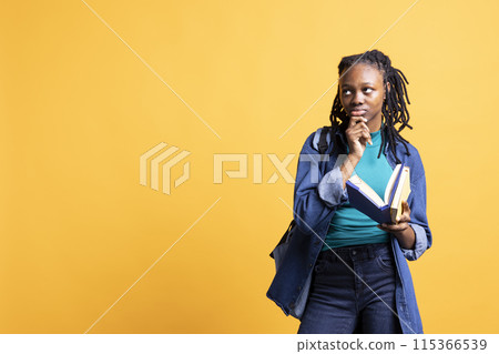 African american woman with book in hands deep in thoughts, isolated over yellow studio background. Bookworm holding novel, thinking about themes and characters after reading 115366539