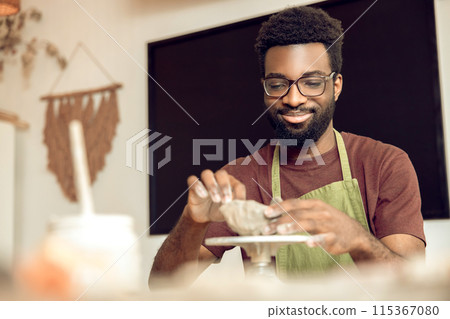 Dark-skinned man in eyeglasses shaping mug from clay and looking focused 115367080