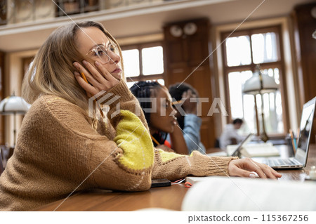 Blonde young woman sitting at the table in the library and feeling tired Blonde young woman sitting at the table in the library and feeling tired 115367256
