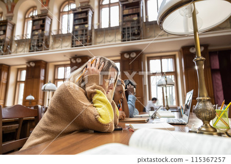 Blonde young woman sitting at the table in the library and feeling tired Blonde young woman sitting at the table in the library and feeling tired 115367257