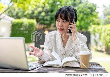 A focused Asian woman is talking on the phone with her colleague while working remotely in a garden. A focused Asian woman is talking on the phone with her colleague while working remotely in a garden. 115370505