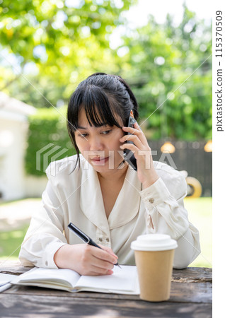 A focused Asian woman is talking on the phone with her colleague while working remotely in a garden. 115370509