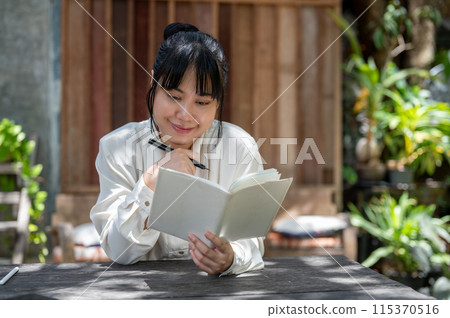 A young positive Asian woman is reading a book or keeping her diary at a table in a garden. A young positive Asian woman is reading a book or keeping her diary at a table in a garden. 115370516