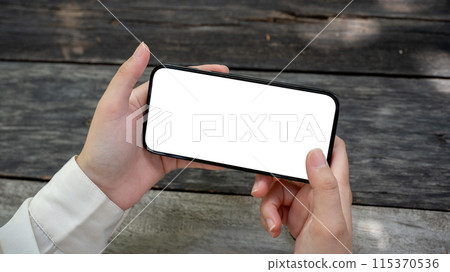 A woman's hand holding a smartphone in a horizontal position over a wooden table in a garden. A woman's hand holding a smartphone in a horizontal position over a wooden table in a garden. 115370536