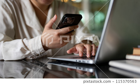 A cropped image of a businesswoman is multitasking, using her smartphone and laptop computer. A cropped image of a businesswoman is multitasking, using her smartphone and laptop computer. 115370563