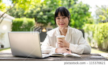 A charming young Asian businesswoman sits at a wooden table in a garden, smiling at the camera. 115370621